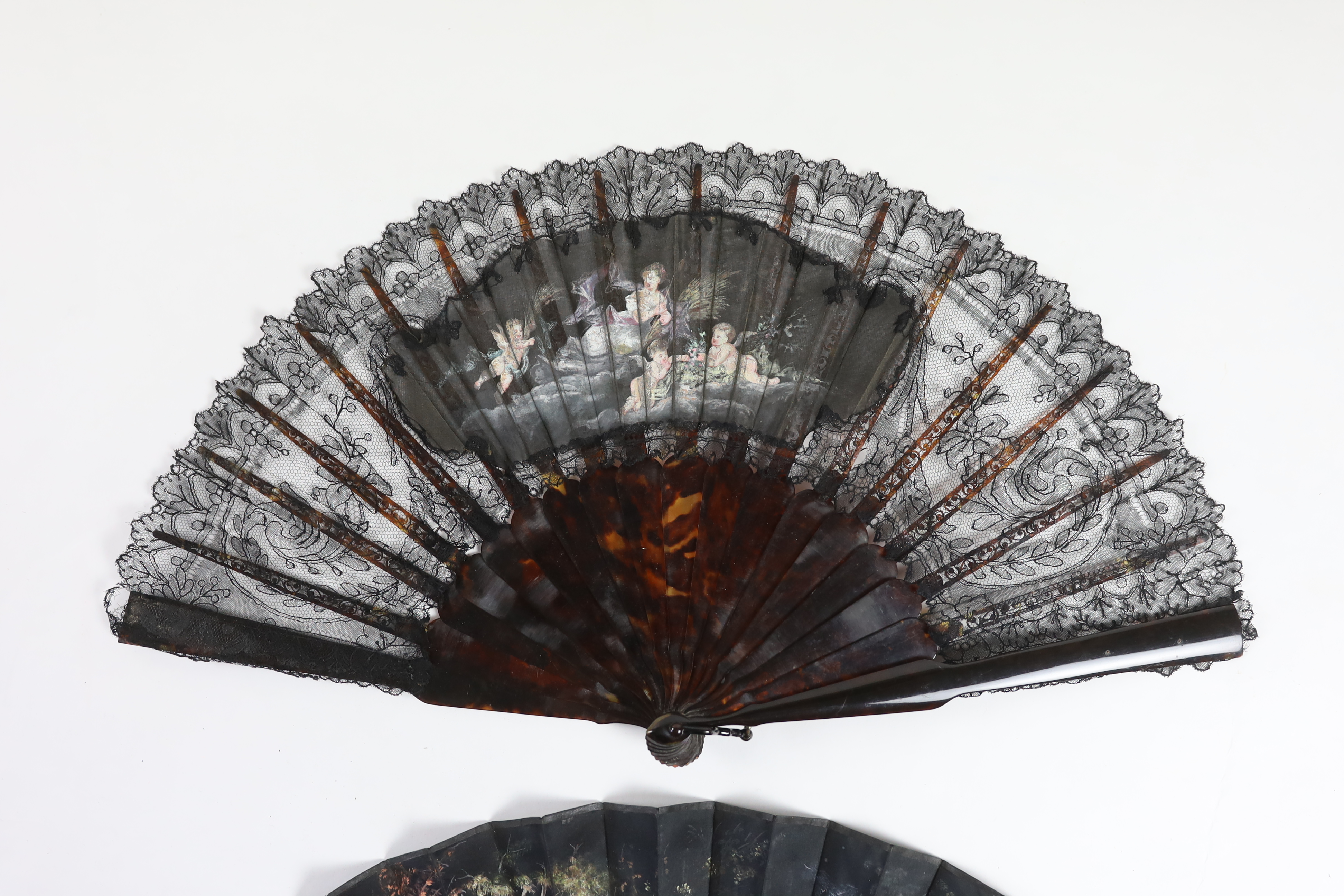 A late Victorian Chantilly black lace and tortoiseshell fan with a central painted cartouche and a mother of pearl and hand painted fairy and cherub fan, indistinctly signed